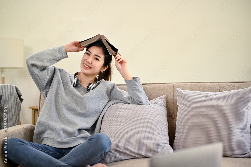 Obraz premium Charming Asian female sits on her comfortable sofa in living room, posing with a book.