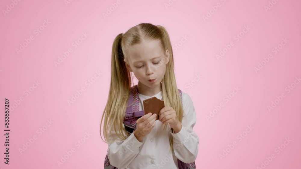 Adorable little schoolgirl with backpack biting chocolate bar, enjoying sweet snack for lunch and smiling to camera Adorable little schoolgirl with backpack biting chocolate bar, enjoying sweet snack for lunch and smiling to camera