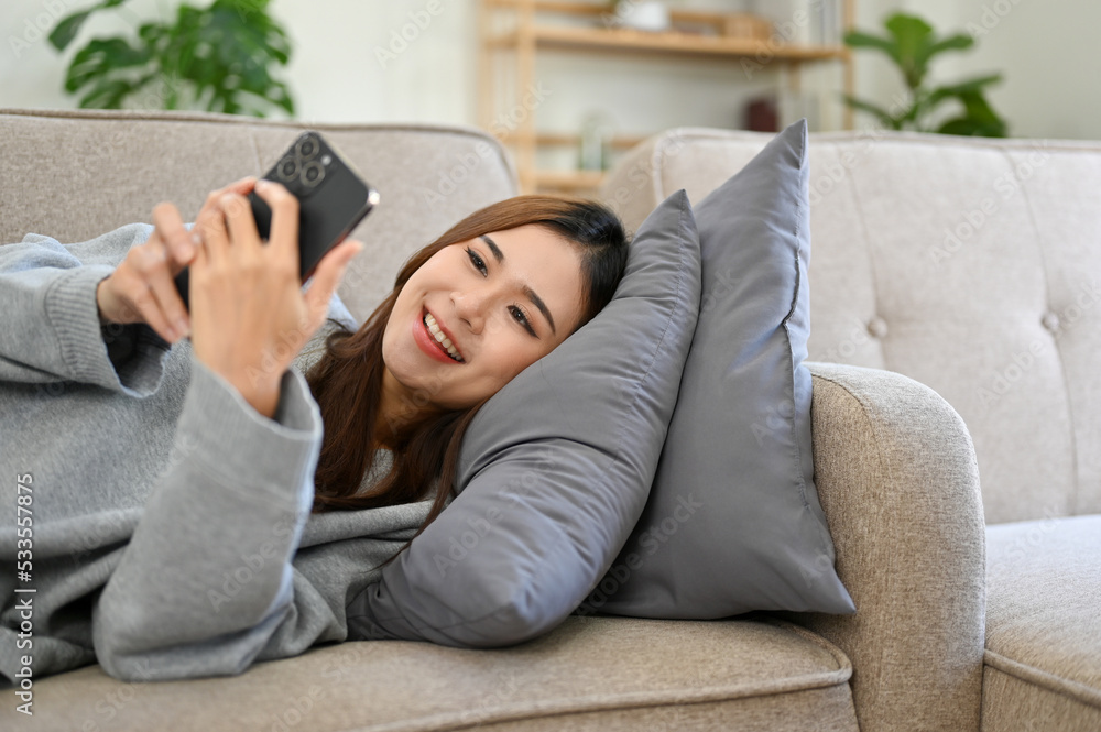Obraz premium Relaxed and happy Asian female using smartphone while laying on her comfortable sofa