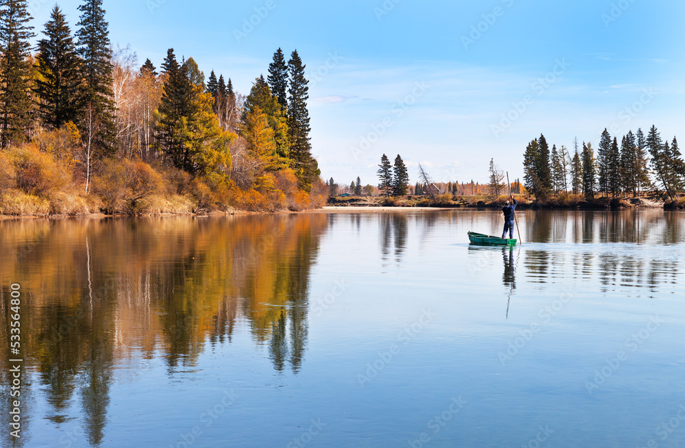 Autumn calm landscape with yellowed trees on the banks of the Irkut River. A village fisherman on a boat returns from fishing. Buryatia, Tunka valley, Nugan village