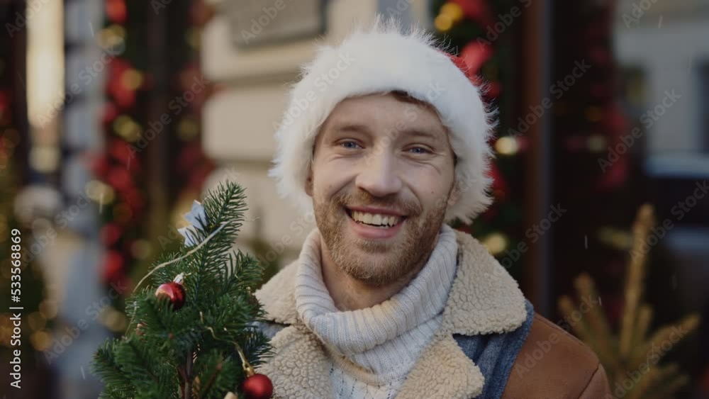 Portrait of the smiled caucasian man wearing santa claus hat standing looking on the camera holding the christmas tree on the blurred decorated street. Close up. People and christmas holiday concept Portrait of the smiled caucasian man wearing santa claus hat standing looking on the camera holding the christmas tree on the blurred decorated street. Close up. People and christmas holiday concept
