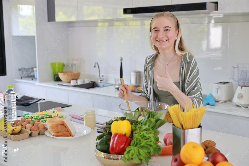 Happy smiling young caucasian girl in casual dress having good time with cooking and making salad on table while showing thumb up in kitchen room at home. Happiness and healthy lifestyle concept.