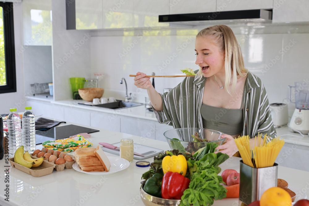 Happy smiling young caucasian girl in casual dress having good time with cooking and making salad on table while tasting food in kitchen room at home. Happiness and healthy lifestyle concept.