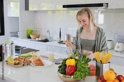 Happy smiling young caucasian girl in casual dress having good time with cooking and making salad on table in kitchen room at home. Happiness and healthy lifestyle concept.