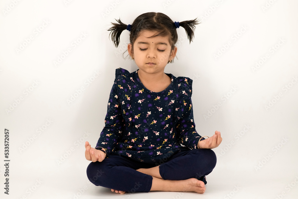 Charming little girl doing serious yoga meditation in studio with an isolated white background. Little Indian girl kid doing meditation in studio.