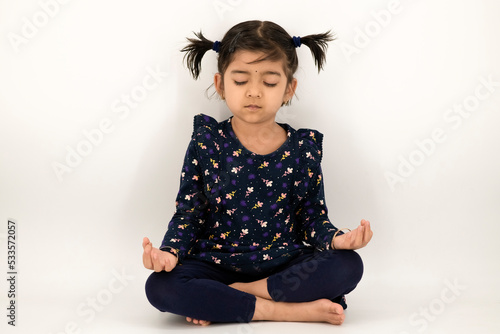Obraz na płótnie Charming little girl doing serious yoga meditation in studio with an isolated white background. Little Indian girl kid doing meditation in studio. w salonie