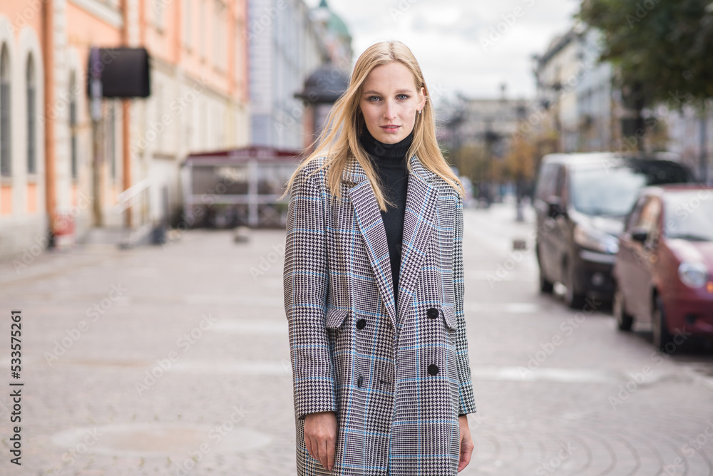 Fototapeta premium Portrait of a young and elegant Caucasian blonde girl in autumn in the city. The concept of femininity and style. A pretty girl is happy to pose in the city on the street in a coat.