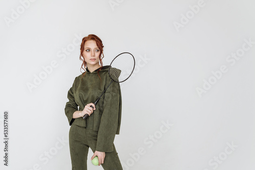 attractive young red-haired woman in a green tracksuit holding a tennis racket on a white background copy space