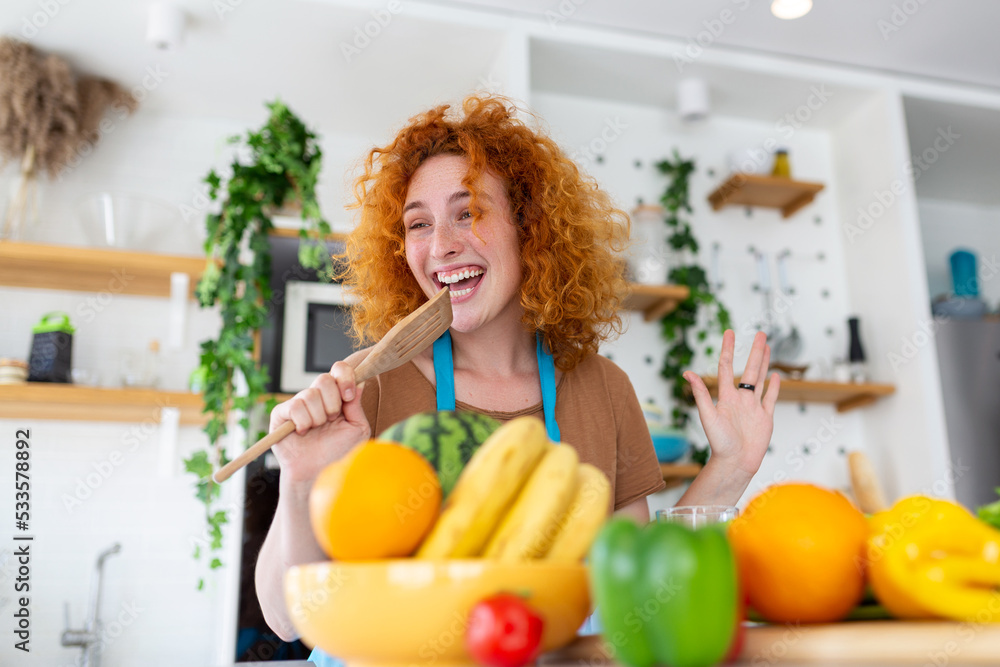 Funny beautiful woman singing into spatula, cooking in modern kitchen, holding spatula as microphone, dancing, listening to music, playful girl having fun with kitchenware, preparing food.
