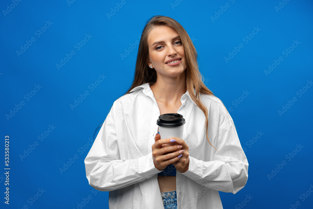 Beautiful attractive young lady holding takeaway paper coffee cup against blue background in studio Beautiful attractive young lady holding takeaway paper coffee cup against blue background in studio