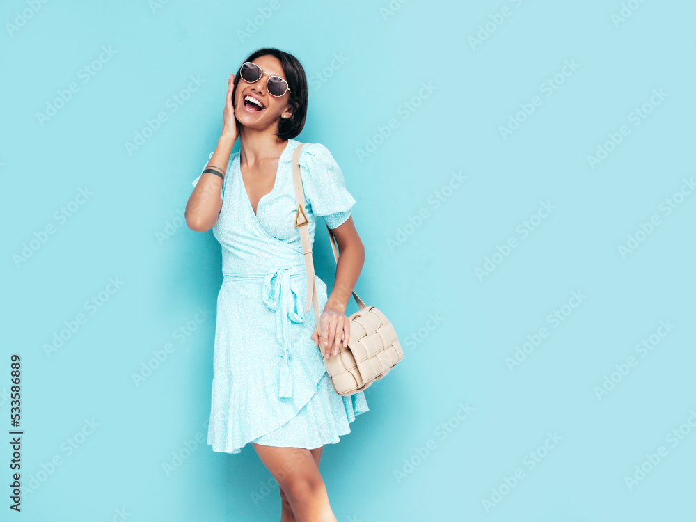 Portrait of young beautiful smiling female in trendy summer dress. Carefree woman posing near blue wall in studio. Positive model having fun indoors with handbag. Cheerful and happy. Isolated Portrait of young beautiful smiling female in trendy summer dress. Carefree woman posing near blue wall in studio. Positive model having fun indoors with handbag. Cheerful and happy. Isolated