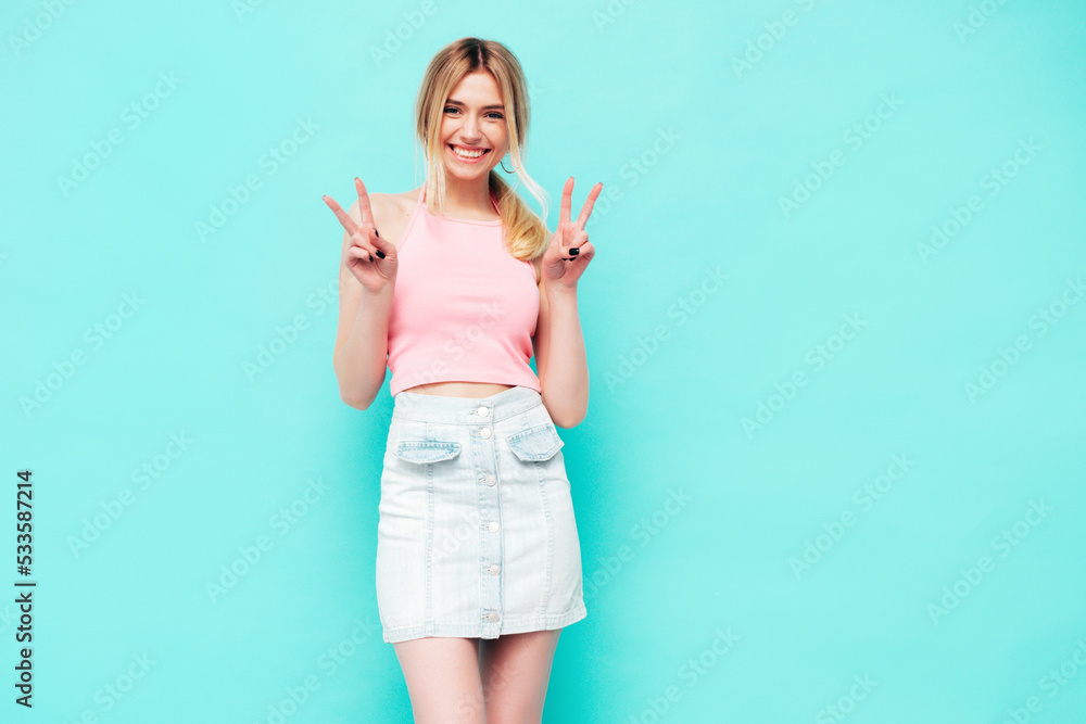 Young beautiful smiling blond female in trendy summer jeans skirt. carefree woman posing near blue wall in studio. Positive model having fun indoors. Cheerful and happy. Shows peace sign Young beautiful smiling blond female in trendy summer jeans skirt. carefree woman posing near blue wall in studio. Positive model having fun indoors. Cheerful and happy. Shows peace sign