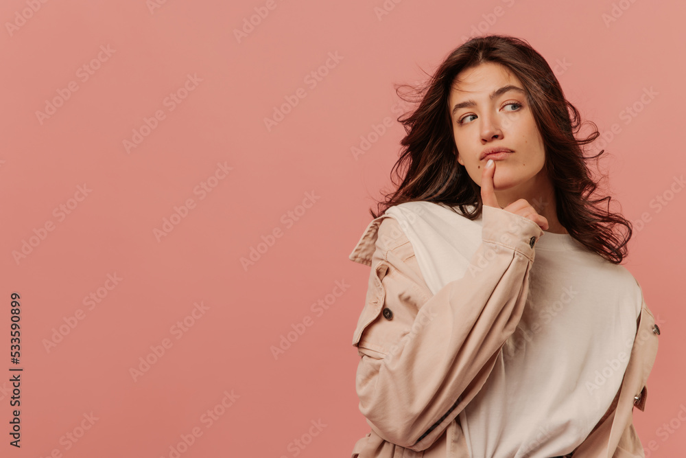 Young caucasian girl looks pensively sideways with her finger on chin against pink background with place for text. Brunette girl with wavy hair wears light-colored casual clothes. Young caucasian girl looks pensively sideways with her finger on chin against pink background with place for text. Brunette girl with wavy hair wears light-colored casual clothes.