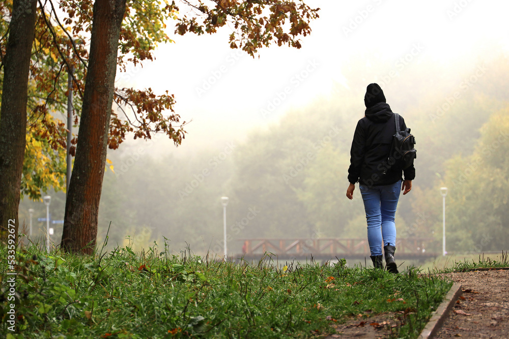 Fog in city park, girl walking on background of autumn trees and lake bridge in morning mist