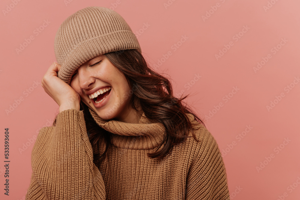 Positive young caucasian girl closes eyes and smiles with teeth pulling hat over face on pink background. Brunette with wavy hair is wearing brown sweater. Lifestyle, feminine beauty concept Positive young caucasian girl closes eyes and smiles with teeth pulling hat over face on pink background. Brunette with wavy hair is wearing brown sweater. Lifestyle, feminine beauty concept