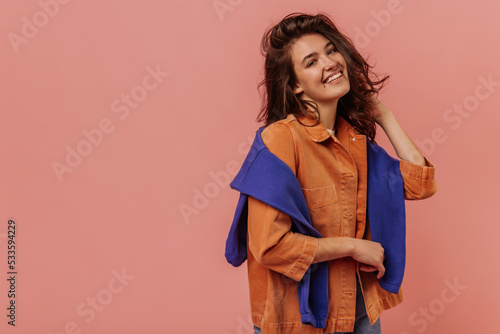 Smiling young caucasian girl with dark, wavy hair looks at camera against pink background. Woman is wearing casual spring clothes. Leisure lifestyle and beauty concept.