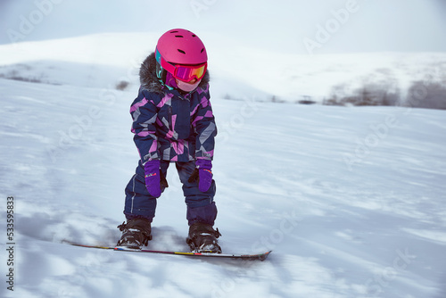 Little girl snowboarder on empty track at ski resort in sunny winter day. Portrait of kid in sportswear with equipment ride on slope
