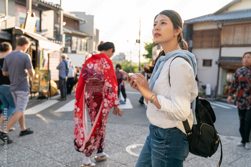 asian chinese female traveler consulting online guide on phone while visiting Ninenzaka and Sannenzaka in Kyoto japan. a girl is wearing red kimono traditional Japanese clothes at background