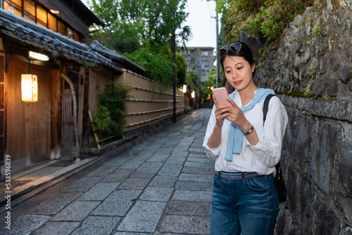 leisure asian Japanese woman tourist checking online for geisha house near her on phone against rock wall on stone pavement alley near Gion Hanamikoji Street in Kyoto japan in the evening photo