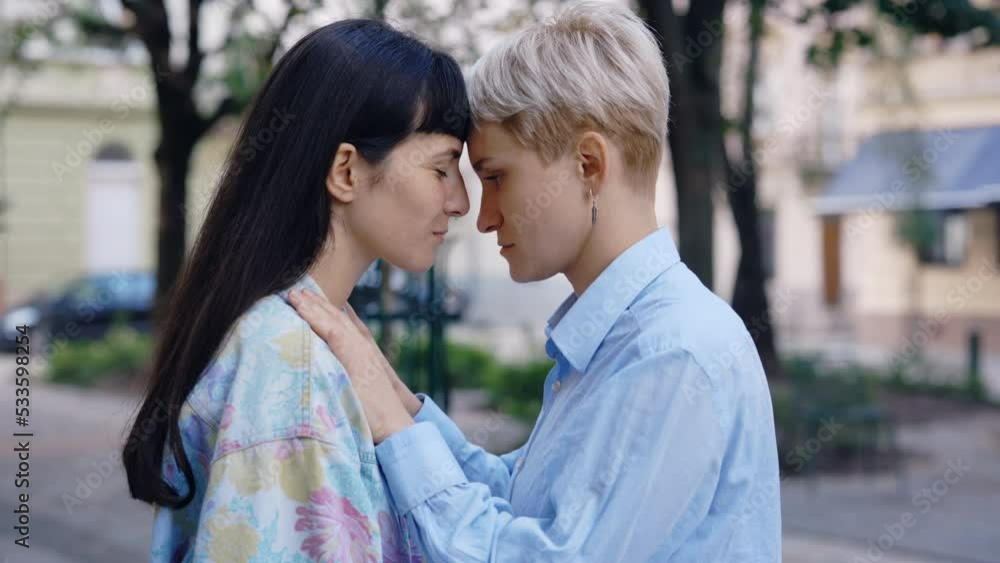 Two lesbian woman, standing in the city centre, one partner kissing her girlfriend in the forehead. Lesbian couple feeling in love together. Romantic date, love, tender relationship Two lesbian woman, standing in the city centre, one partner kissing her girlfriend in the forehead. Lesbian couple feeling in love together. Romantic date, love, tender relationship