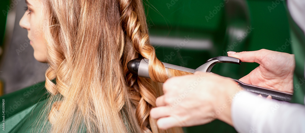 Fototapeta premium The female hairdresser is curling hair for a brown-haired young caucasian woman in a beauty salon