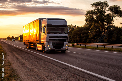Foto A semitrailer tractor with a tilt semitrailer transports cargo against the backdrop of an evening sunny sunset in summer