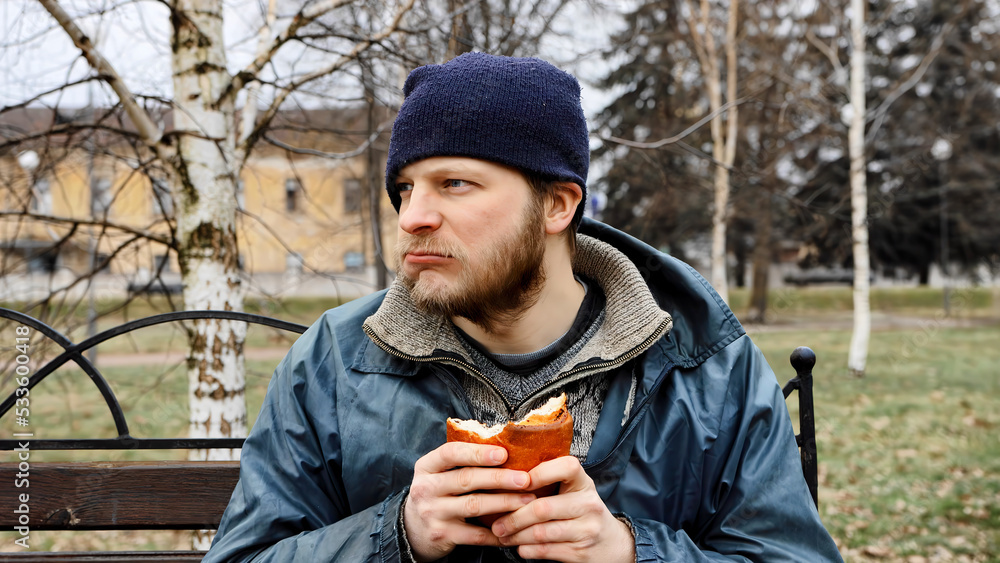 Hungry bearded homeless man sits on a bench and eats a bun in a city park. Below poverty line. Male tramp in dirty clothes with cap and hat. Beggar. Illegal immigrant. Drunkenness, alcoholism Hungry bearded homeless man sits on a bench and eats a bun in a city park. Below poverty line. Male tramp in dirty clothes with cap and hat. Beggar. Illegal immigrant. Drunkenness, alcoholism