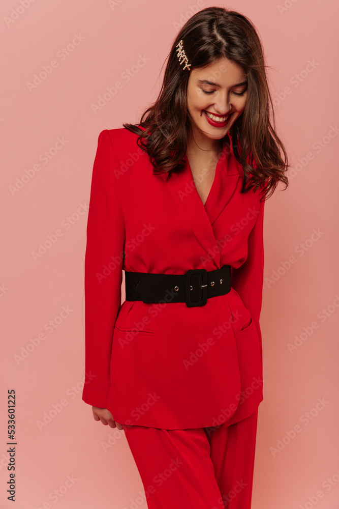 Pretty caucasian young dark-haired woman with smile on her face looks down on pink background. Model wears glamorous red suit with black belt. Lifestyle, different emotions, leisure concept. Pretty caucasian young dark-haired woman with smile on her face looks down on pink background. Model wears glamorous red suit with black belt. Lifestyle, different emotions, leisure concept.