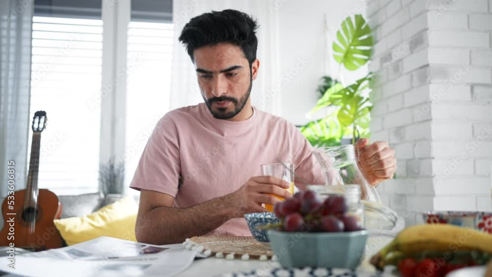 Young man pouring orange juice in glass and reading newspaper, morning routine concept. Young man pouring orange juice in glass and reading newspaper, morning routine concept.