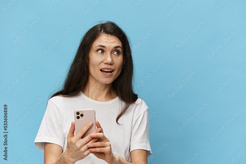 a happy, joyful woman with beautiful long hair, in a light T-shirt, stands with a fashionable phone in her hands. Horizontal photo on a light blue background with space for text a happy, joyful woman with beautiful long hair, in a light T-shirt, stands with a fashionable phone in her hands. Horizontal photo on a light blue background with space for text