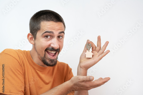 Happy young man smiling with the new home keys. Keychain in the shape of a wooden house on white background.