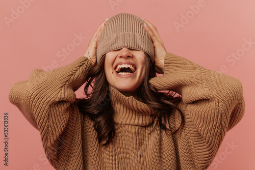 Cheerful young caucasian girl is laughing holding hat on head, pulled over eyes on pink background. Brunette girl is wearing everyday warm clothes. People's emotions, lifestyle and fashion concept. 