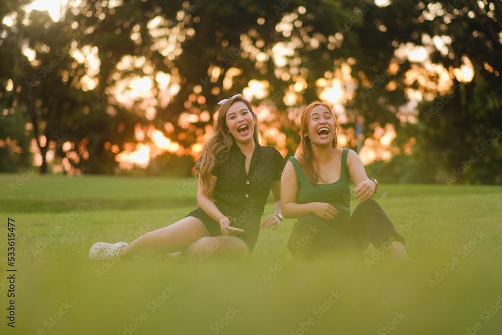 Naklejka premium Two young Asian women, both with long wavy hair, howl with laughter. They are sitting beside each other on the grass on a beautiful summer day at sunset.