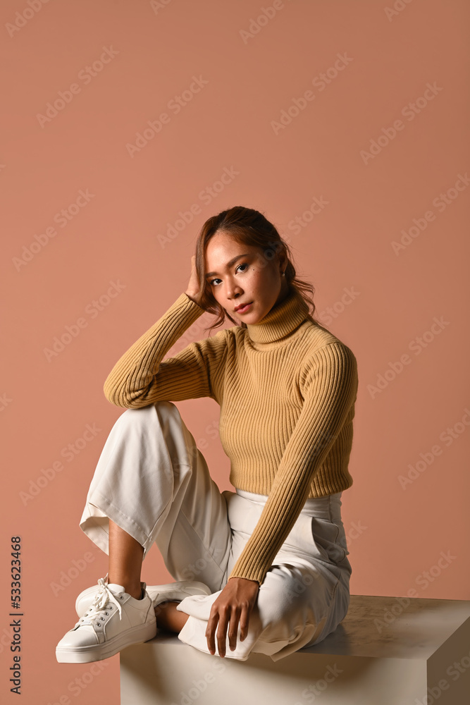 Fototapeta premium Portrait of trendy woman wearing warm sweater sitting on pink background. Fashion studio photo, Autumn and Winter concept