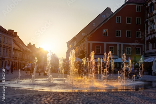 Flowing fountain in center of Maribor in golden hour. Splashing water on square in old city in Slovenia in sunset light. Historical building in turistic town.