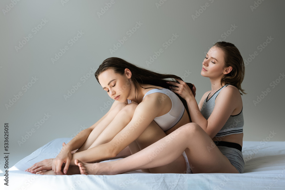 young lesbian woman touching long hair of brunette girlfriend sitting on bed with closed eyes on grey background. young lesbian woman touching long hair of brunette girlfriend sitting on bed with closed eyes on grey background.