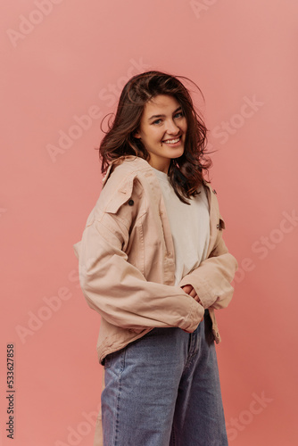 Stylish young brunette caucasian woman is smiling looking at camera on pink background. Hottie wears white shirt, beige jacket and jeans. Lifestyle concept 