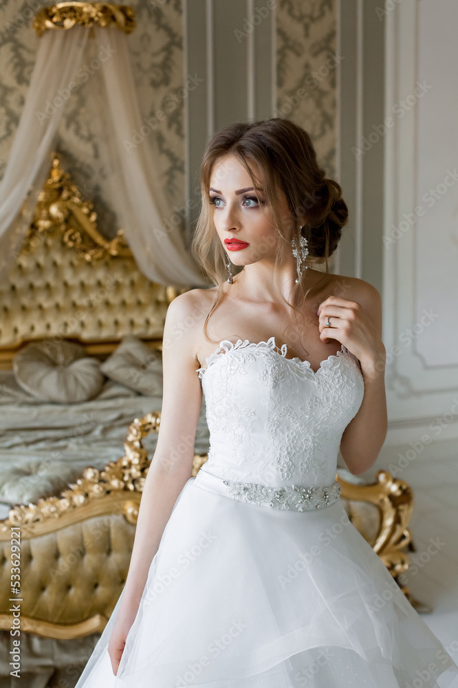 A beautiful smiling bride with chic makeup and hairstyle in a white wedding dress is standing in a bedroom with a stylish light vintage interior, in the morning before the wedding. A beautiful smiling bride with chic makeup and hairstyle in a white wedding dress is standing in a bedroom with a stylish light vintage interior, in the morning before the wedding.