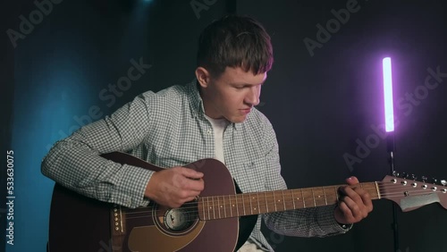 A man plays guitar in a dark studio with bright colors. A young guy is practicing the guitar, learning new chords and notes. . High quality 4k footage