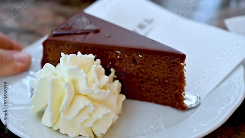 Vienna, Austria, August 2022. Nice slow motion footage of a Viennese specialty: a nice slice of sachertorte served on a plate with some whipped cream next to it. The hand approaches the plate.