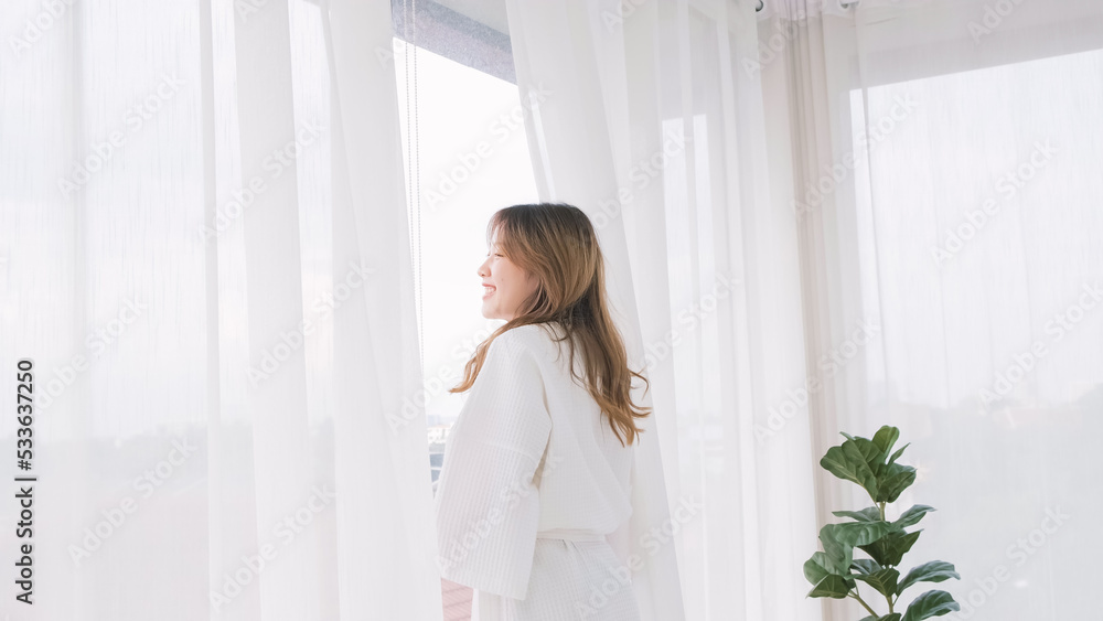 Woman wearing bathrobe opens curtains in bedroom and see around the outdoors. Welcome new sunny day, happy positive young woman wakes up and looks in the window at home or hotel feeling optimistic. Woman wearing bathrobe opens curtains in bedroom and see around the outdoors. Welcome new sunny day, happy positive young woman wakes up and looks in the window at home or hotel feeling optimistic.