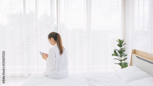 The back of long tied hair Woman in a bathrobe using a smartphone while sitting on the bed with a closed white curtain over the glass window in the background. Image with copy space.