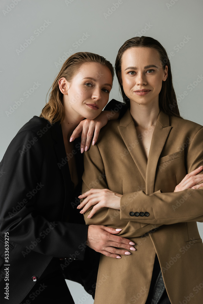 young woman leaning on lesbian girlfriend standing with crossed arms and looking at camera isolated on grey.