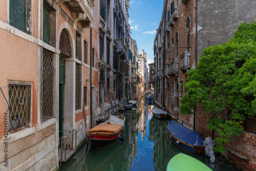 Fototapeta Naklejka Na Ścianę i Meble -  Boats near vintage brick walls of houses on the water surface of a narrow canal street in Venice, reflections and sky on a Venetian street, a tree on the bank of a Venice canal on a sunny day
