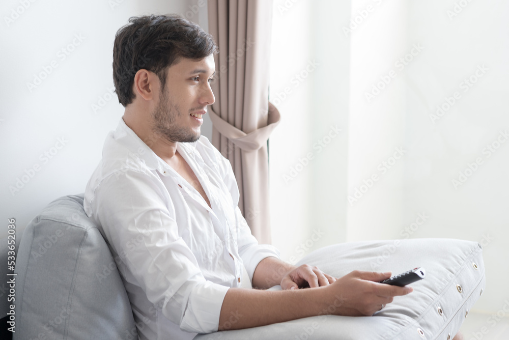 Portrait of a happy young man sits on comfortable sofa holds remote control and watche television at home in living room Portrait of a happy young man sits on comfortable sofa holds remote control and watche television at home in living room