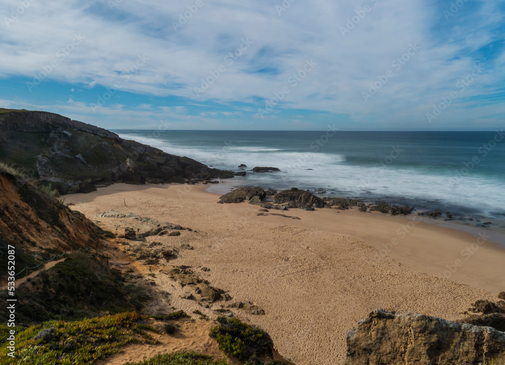 View of empty small sand beach with with long exposure blurred ocean waves and sharp rock and cllifs at wild Rota Vicentina coast near Porto Covo, Portugal.