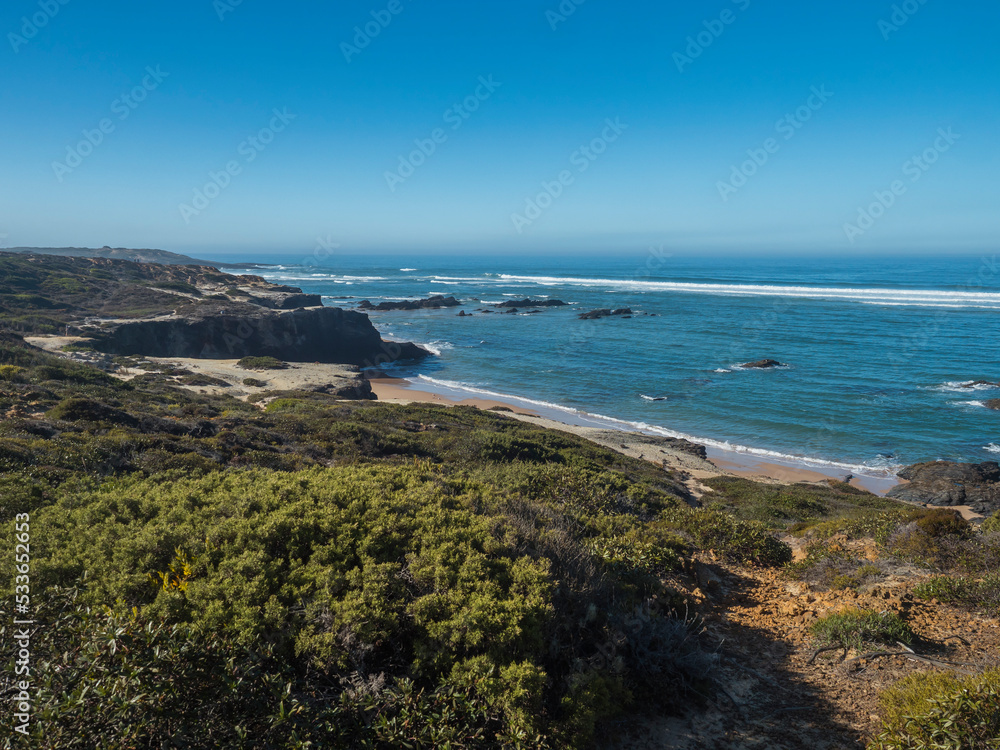 View of Praia das Furnas golden sand beach with ocean waves and sharp rock and green vegetation at wild Rota Vicentina coast near near Vila Nova de Milfontes, Portugal.