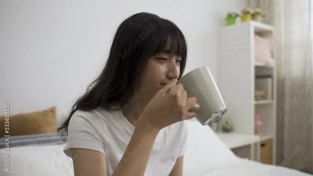 cheerful asian girl smelling and sipping morning tea from cup and nodding head feeling contented with the good quality in the bedroom at home. cheerful asian girl smelling and sipping morning tea from cup and nodding head feeling contented with the good quality in the bedroom at home.