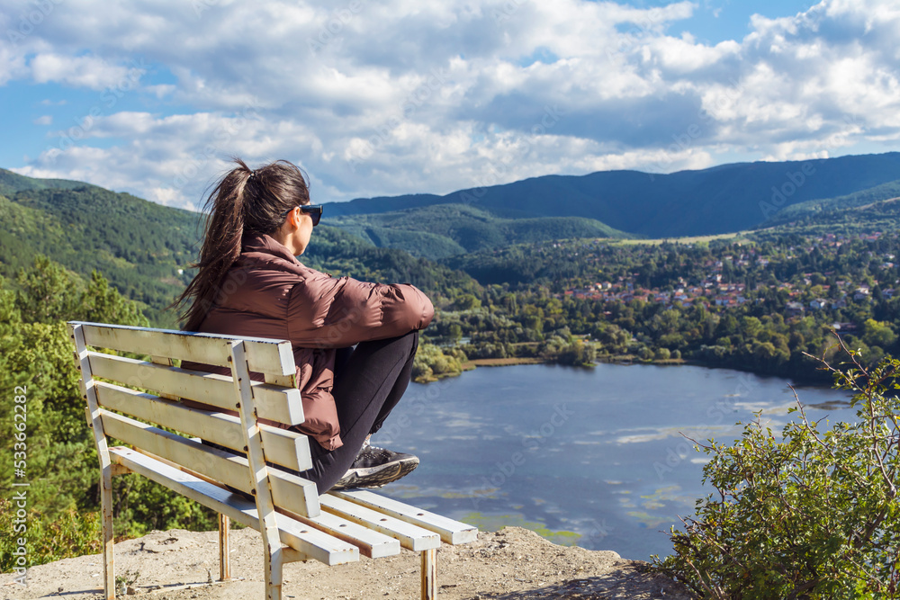 Traveler woman sitting on a bench high above a blue lake with stunning view . Scenic landscape from Bulgaria ,Pancharevo lake 