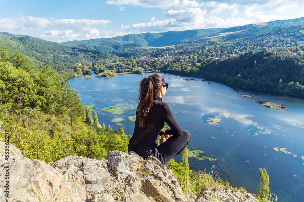 Naklejka premium Traveler woman sitting on a rock high above a blue lake with stunning panoramic view .Traveling in the nature in Bulgaria . Pancharevo Lake